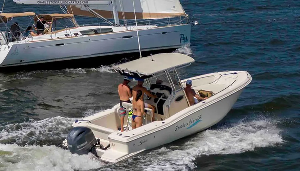 A group of people are enjoying a sunny day on a small white motorboat on the water with a larger sailboat visible in the background