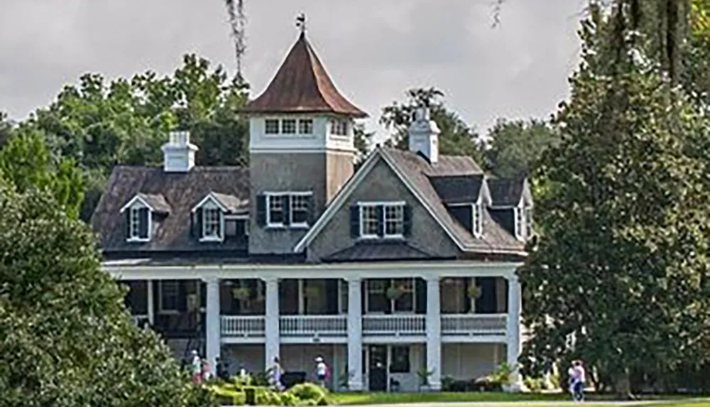 The image shows a large stately historic house with multiple stories dormer windows and a prominent tower set in a lush landscape with people visible in the foreground