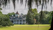 An elegant two-story house with a conical roof feature is nestled among large trees draped with Spanish moss, overlooking a well-manicured lawn.