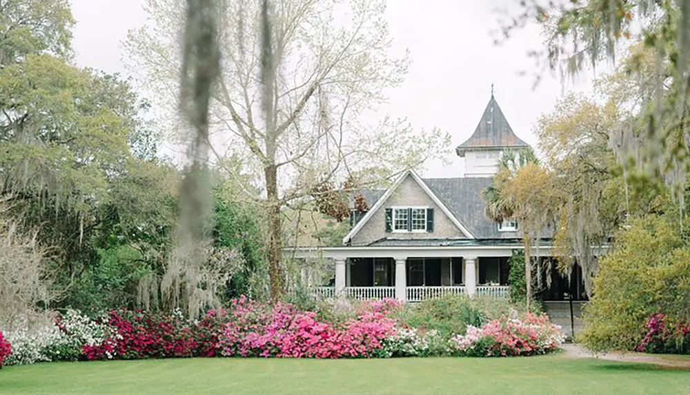 The image shows a charming house with a peaked roof and a welcoming porch surrounded by lush gardens and draped with Spanish moss creating an idyllic Southern scene