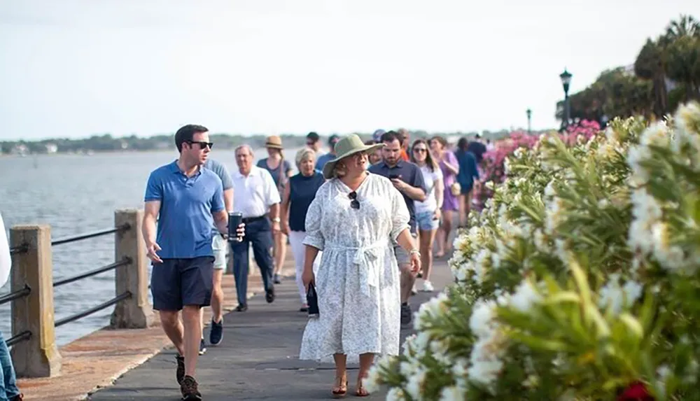 A diverse group of people is enjoying a sunny day along a waterside walkway lined with lush flowering plants