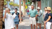 A group of people are attentively listening to a woman gesturing with her hand as if she is guiding a tour on a city street.