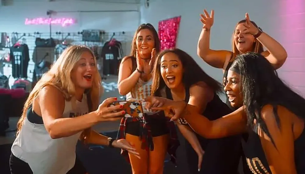 A group of joyful women is gathered around snapping a group selfie with one holding a selfie stick all evidently enjoying the moment and each others company