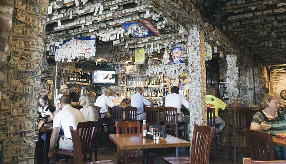 The image shows the interior of a unique bar or restaurant where the walls and ceiling are covered with dollar bills with patrons sitting at tables dining or having drinks