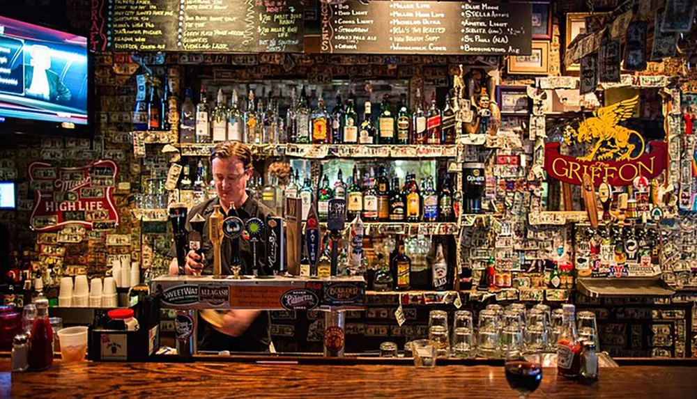 A bartender stands behind a colorful bar adorned with an eclectic assortment of bottles taps and stickers as he pours a drink