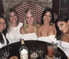 Five women are smiling for a photo at a table with drinks in a social setting