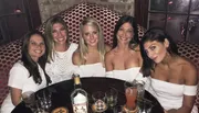 Five women are smiling for a photo at a table with drinks in a social setting.