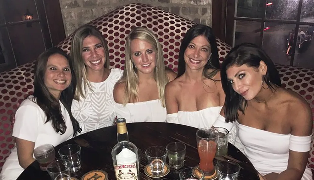 Five women are smiling for a photo at a table with drinks in a social setting