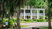 The image shows a classic southern-style manor with large columns, surrounded by a lush garden and Spanish moss-draped trees, where people are leisurely spending time in its tranquil setting.