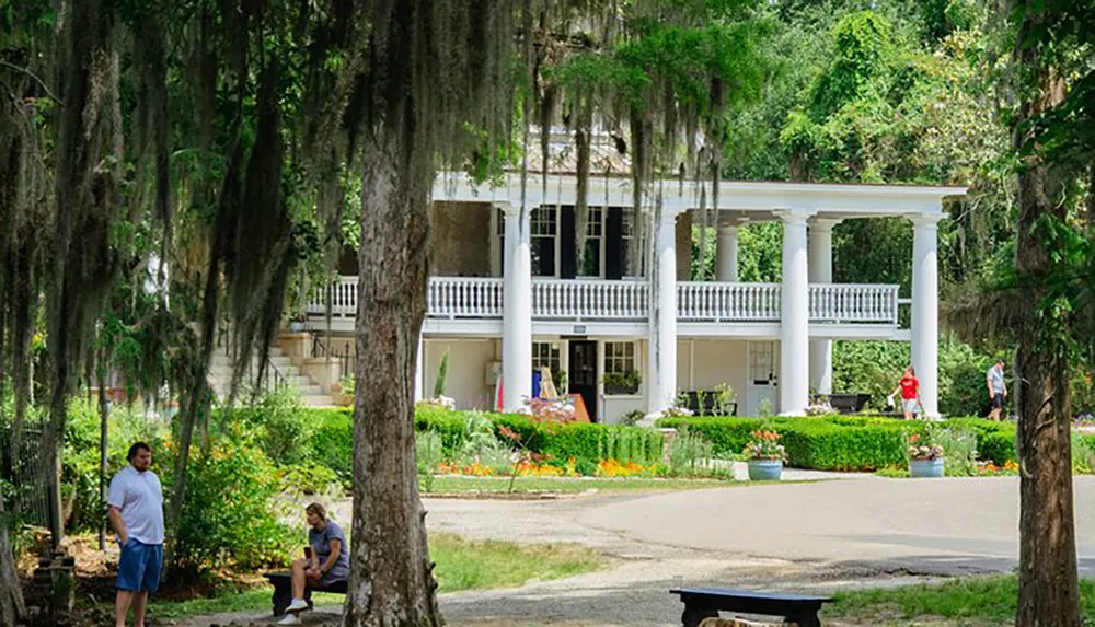 The image shows a classic southern-style manor with large columns surrounded by a lush garden and Spanish moss-draped trees where people are leisurely spending time in its tranquil setting