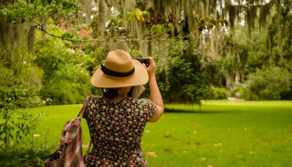 A person in a floral dress and hat is taking a photo in a lush green garden draped with Spanish moss