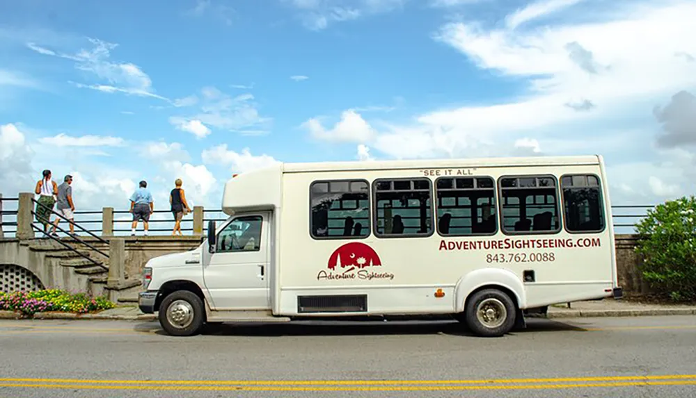 A sightseeing bus with the text Adventure Sightseeing is parked by the side of the road while a group of people walks across a bridge in the background under a partly cloudy sky