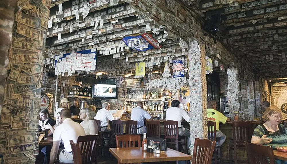 The image shows the interior of a bar or restaurant where the walls and ceiling are adorned with numerous dollar bills and patrons are dining and socializing