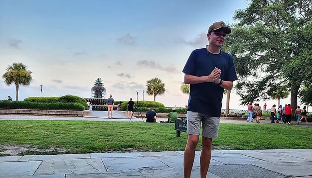 A person stands in the foreground in a park setting with a palm tree and people in the background during what appears to be dusk