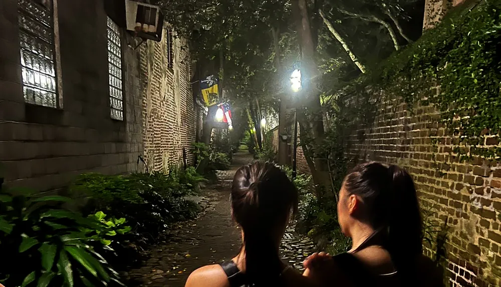 Two individuals are walking away from the camera down a narrow brick-walled alley illuminated by overhead lights at night