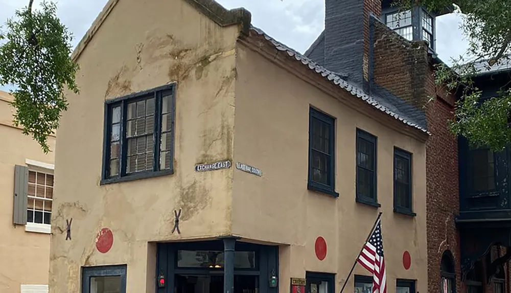 The image shows an old two-story stucco building with a flagstone roof several windows with dark frames a mounted American flag and street signs indicating the corner of Exchange and East Bay streets