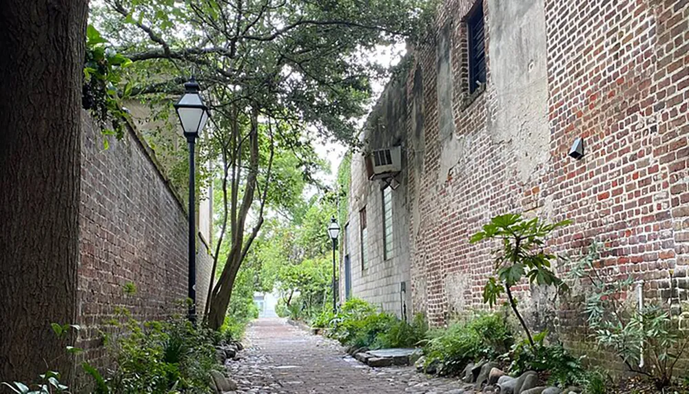 The image shows a cobblestone alleyway lined with vintage-style street lamps and bordered by a brick building on one side and lush greenery on the other