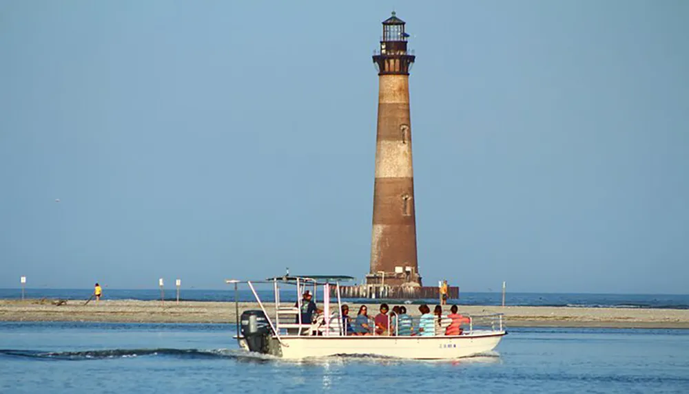 A boat with passengers is sailing near a shoreline with a tall slender lighthouse in the background