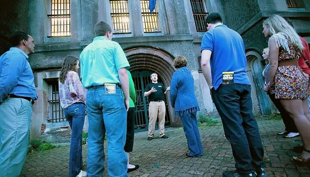 A group of people is listening attentively to a guide who is speaking in front of a historic building with barred windows