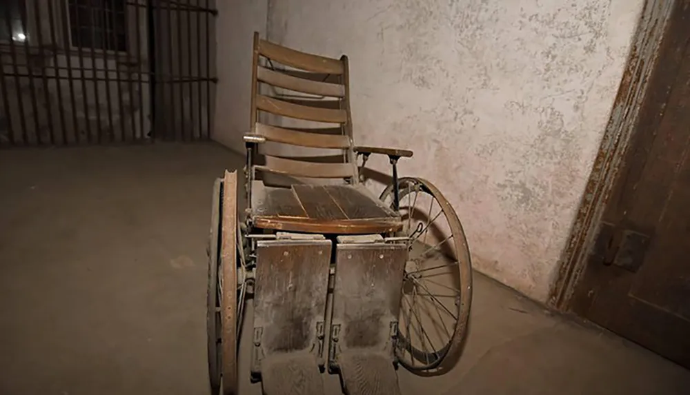 An old weathered wheelchair sits abandoned in a dimly lit room with barren walls and a metal-barred gate in the background