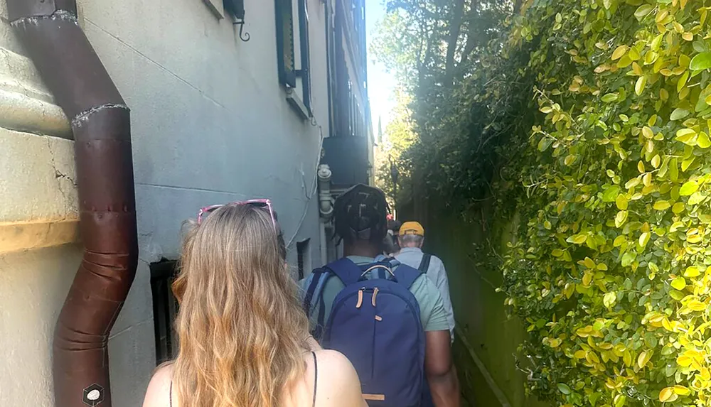 A group of people is walking through a narrow alleyway lined by a building on one side and dense foliage on the other