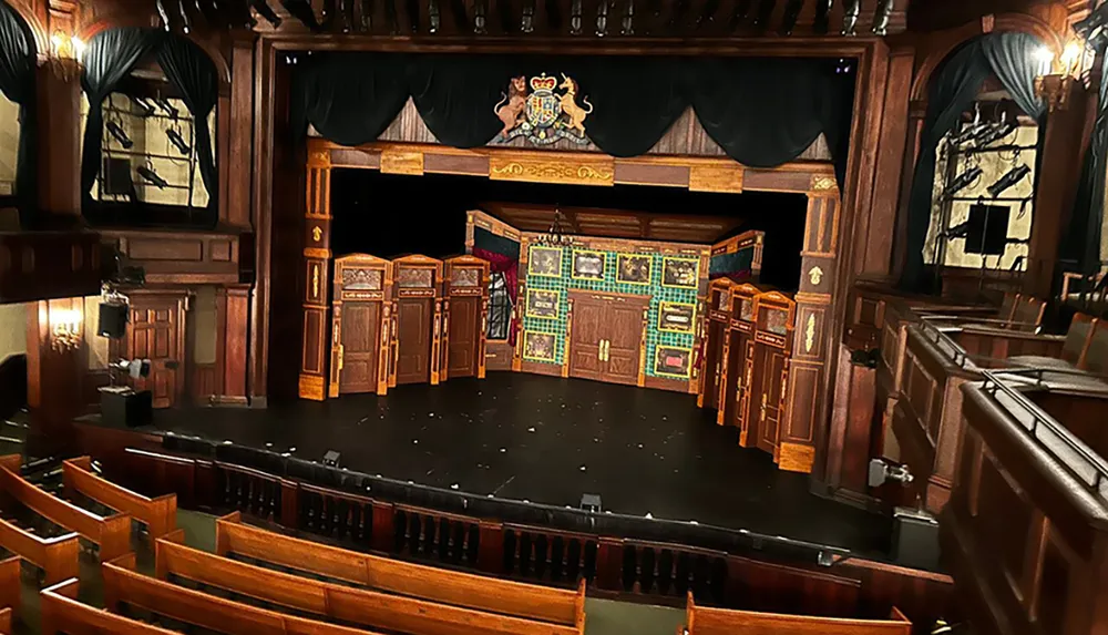 The image shows an ornate empty theater with wooden seating facing a stage adorned with a decorative proscenium and set pieces conveying a sense of anticipation for a performance