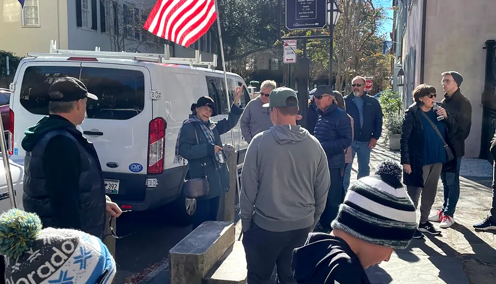 A group of people is gathered around a tour guide near a white van on a sunny street with an American flag visible in the background