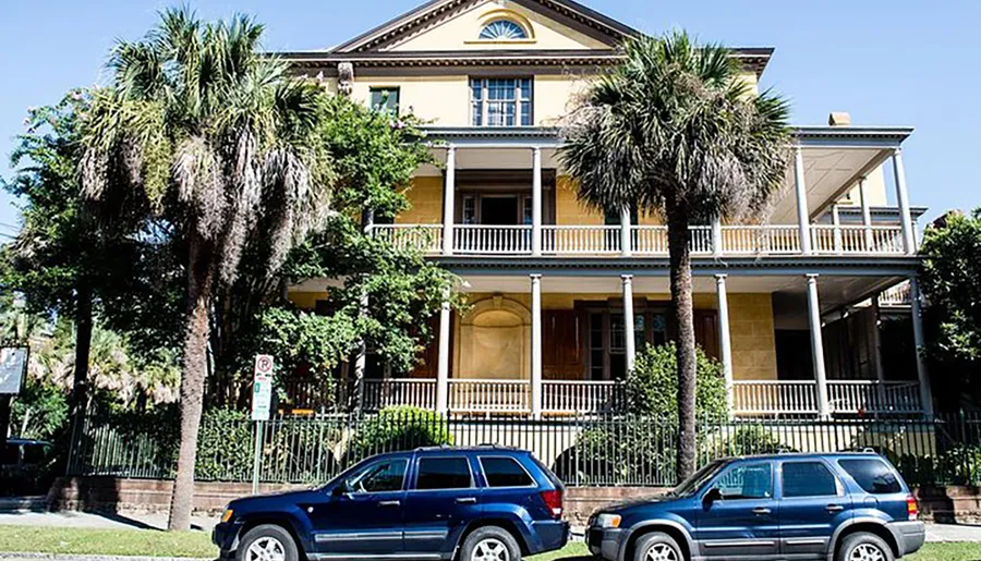 The image depicts a stately two-story yellow house with white trim, balconies, and columned porches, flanked by palm trees, with parked cars in front.