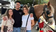 Three smiling people pose for a photo next to a horse that is geared up, possibly for a carriage ride.