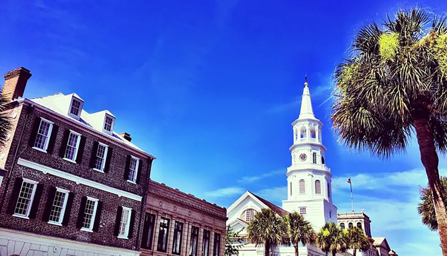 The image features a historic steepled building with a clock tower adjacent to traditional brick buildings and a palm tree under a clear blue sky.