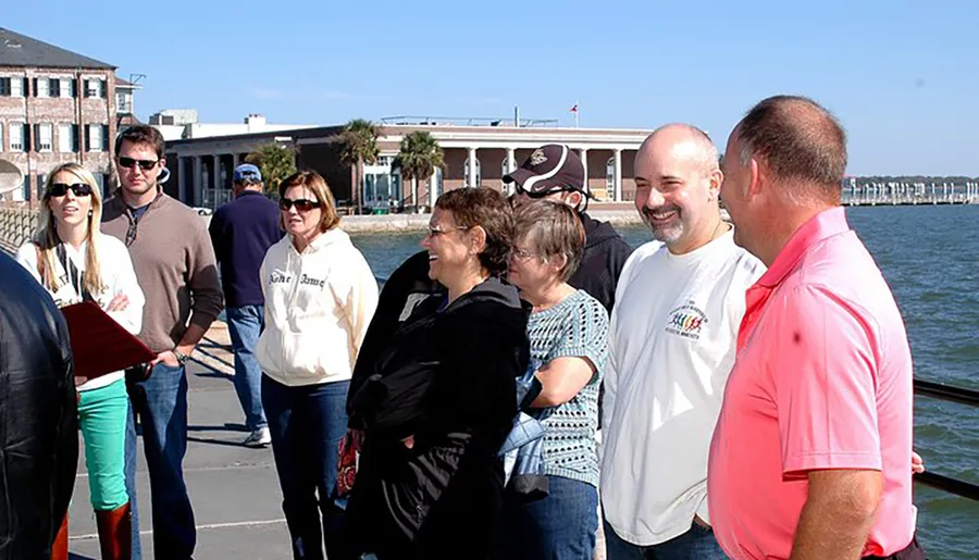 A group of people appears to be enjoying a sunny day at a waterfront location with buildings and a pier in the background.