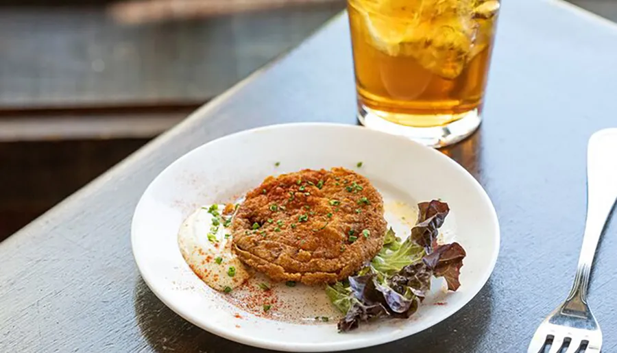 The image shows a breaded and fried food item, possibly a schnitzel or cutlet, served with a dollop of sauce and a side salad, accompanied by a glass of iced tea on a wooden table.