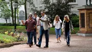 A group of people is walking through a park-like setting with trees and a lamp post, with one man gesturing mid-conversation.