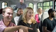 Several people are smiling and enjoying a ride on a shuttle bus, with one man pointing at something out of frame, capturing a moment of shared amusement.