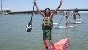 A person is jubilantly raising their arms while kneeboarding, with other individuals stand-up paddleboarding in the background.