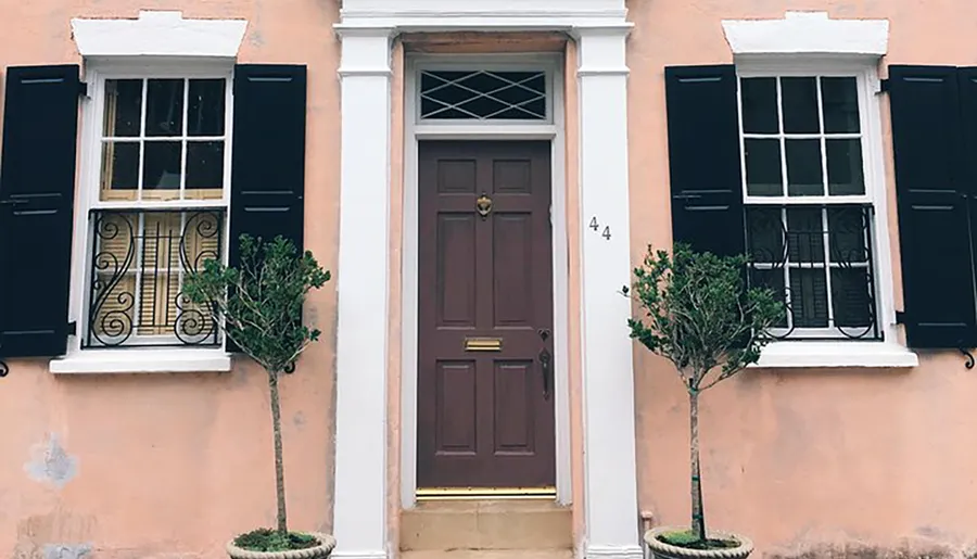 The image shows a symmetrical peach-colored facade with a centered door flanked by two windows with black shutters and two potted plants.