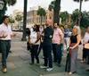 A group of people attentively listens to a man who appears to be giving a tour or a talk outside on a city sidewalk