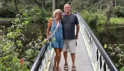 A smiling couple is standing together on a white-railed bridge set in a lush, green environment with a water body on the side.