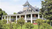 The image shows a grand two-story house with a large wrap-around porch, columns, and a prominent turret, nestled in a lush green setting with people casually gathered out front.