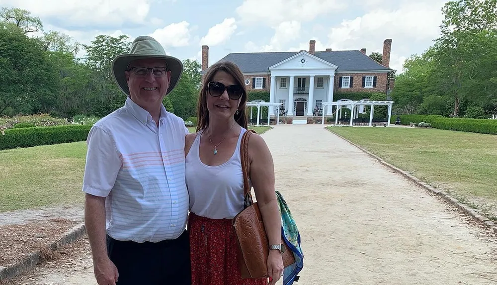 A smiling couple poses in front of a large house with columns and a pathway leading to it suggesting a visit to a historical or significant estate