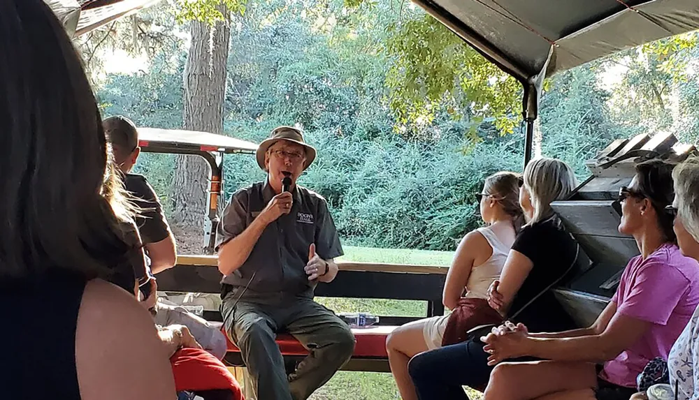 A person wearing a hat is speaking into a microphone while sitting in front of a group of attentive listeners possibly during an outdoor event or tour