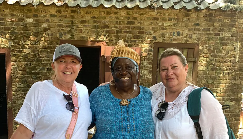 Three smiling women pose together in front of a brick building under a sunny sky