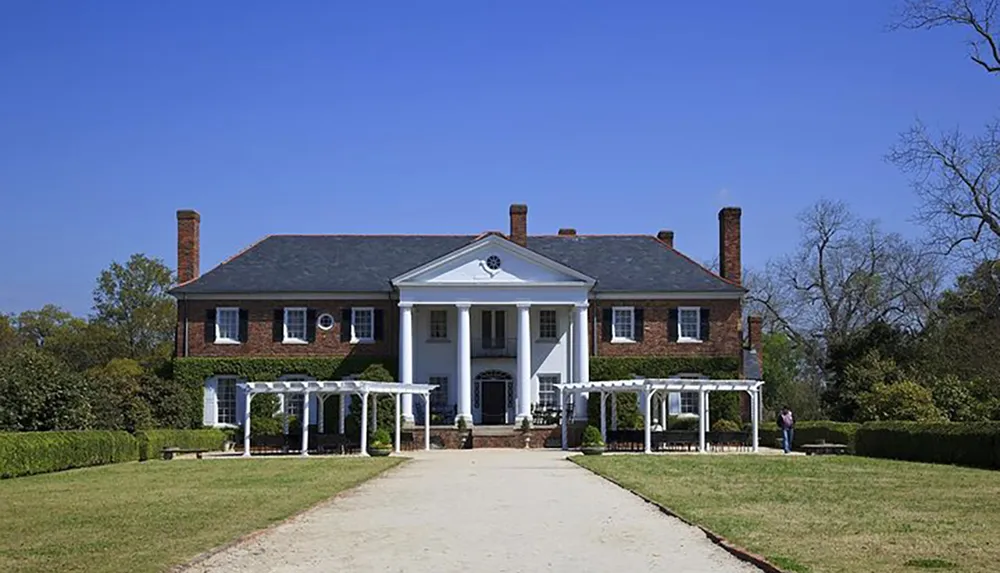 This image shows a symmetrical two-story brick mansion with white columns and window frames surrounded by manicured lawns under a clear blue sky