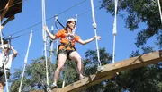 A person wearing a helmet and safety harness is carefully walking across a wooden beam in a ropes course challenge, with trees in the background.