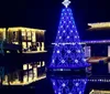 The image depicts a vibrant blue Christmas tree with glowing decorations reflecting on water set against a backdrop of illuminated buildings at night