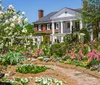 A traditional brick house stands behind a vibrant garden full of blooming flowers and lush greenery under a clear blue sky