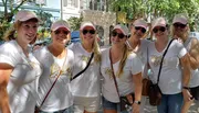 A group of women wearing matching white t-shirts and pink caps, with one wearing a shirt labeled 