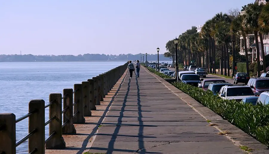 The image shows a peaceful promenade by the water with people walking, a row of parked cars, and tree-lined streets on a sunny day.