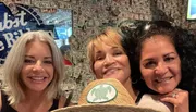 Three smiling women pose together for a photo with a background wall covered in dollar bills.