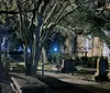 A nighttime view of an old cemetery with weathered gravestones lit by ambient lights that cast shadows among the trees